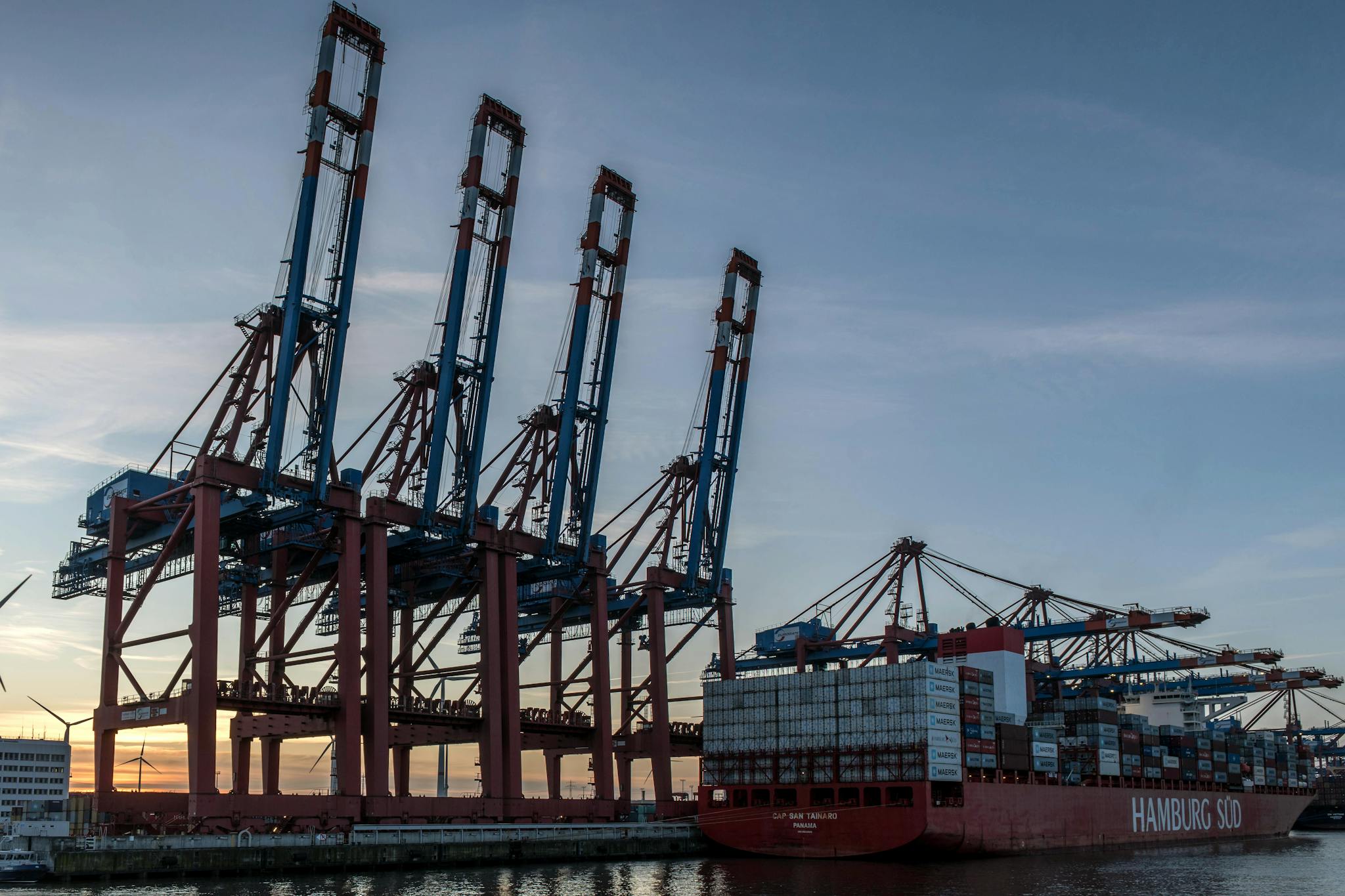 Container cranes and a cargo ship at Hamburg port during sunset, showcasing industrial logistics.